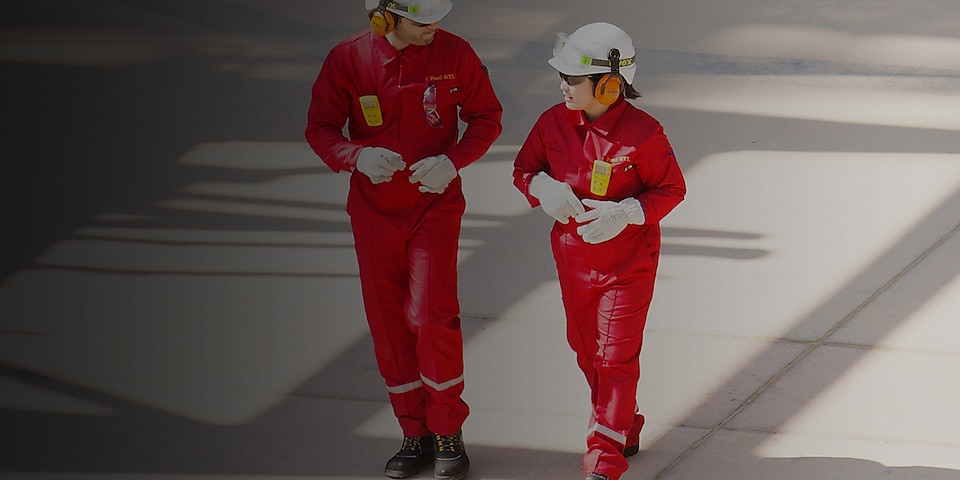 Idący pracownicy i pracownice w czerwonych kombinezonach i białych kaskach ze słuchawkami ochronnymi