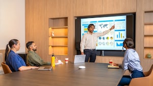 Man giving a presentation in a modern conference room, pointing to a large screen with charts and graphs, while three other people sit at a wooden table with laptops and coffee mugs.​