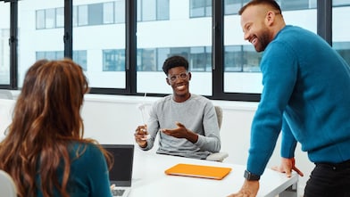 Three people are in the office meeting room with large windows. Man is standing and leaning on the white table, engaging with woman and man seating at this table.​.