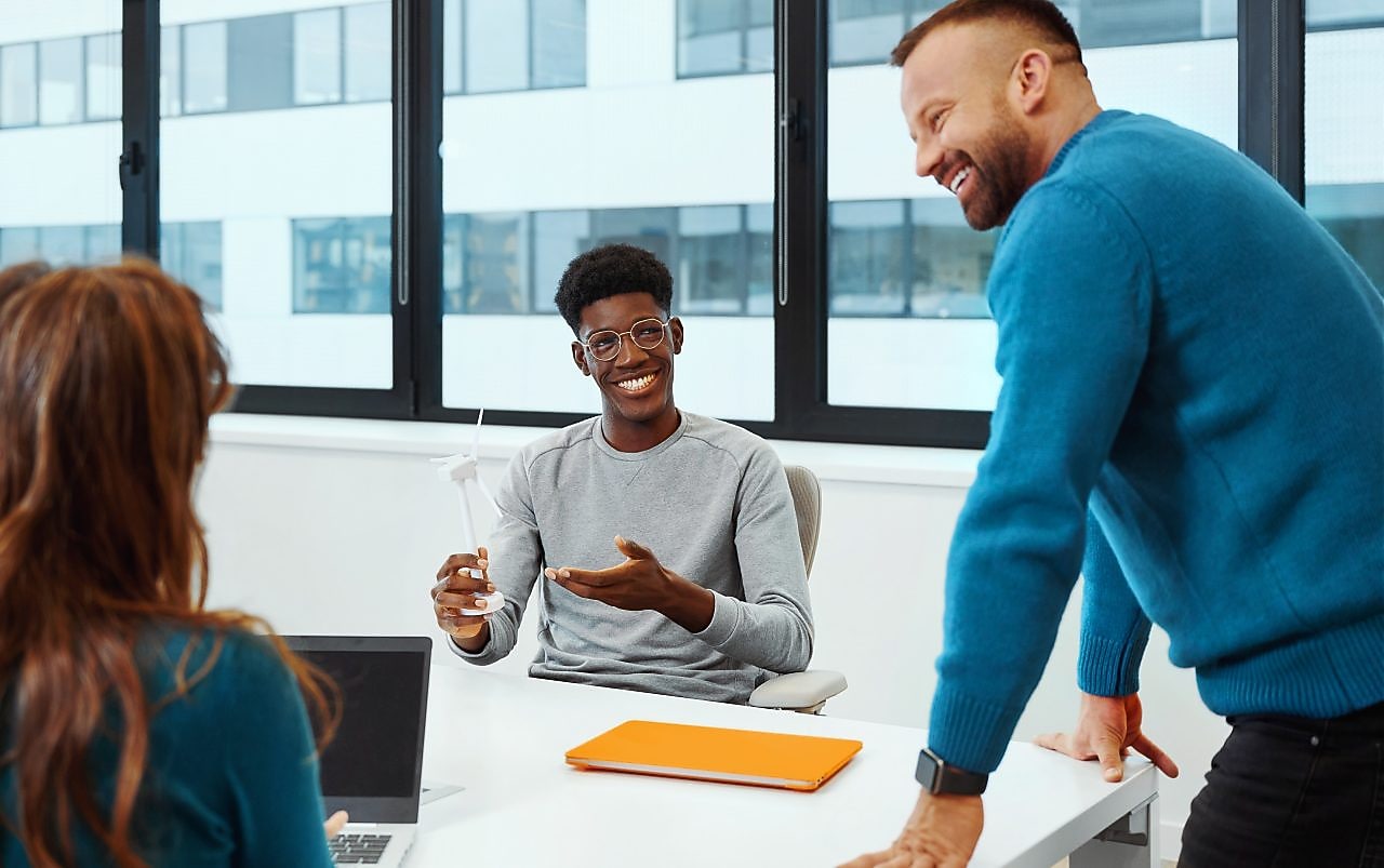 Three people are in the office meeting room with large windows. Man is standing and leaning on the white table, engaging with woman and man seating at this table.​.