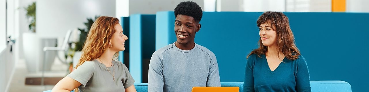 Three people sitting on sofas in a modern indoor space, talking and holding reusable drink containers. There is a large blue partition panel in the background.​