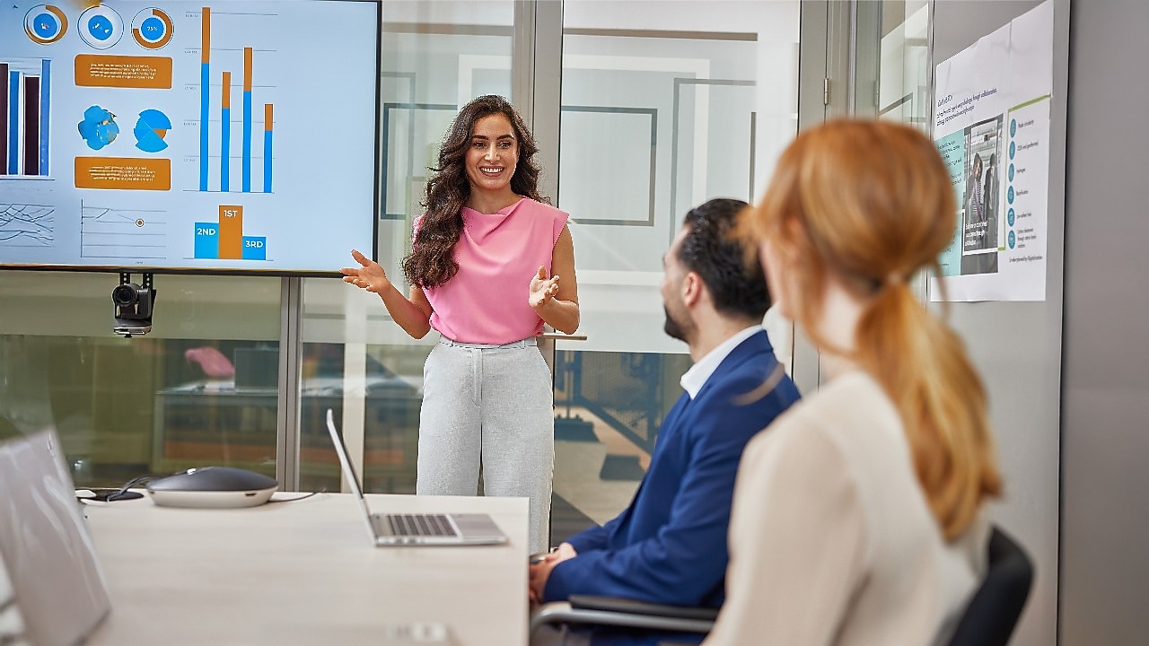 Woman presenting in a meeting room, standing near a screen with colorful charts and graphs, while a man and a woman sit at a table with a laptop and notes.​