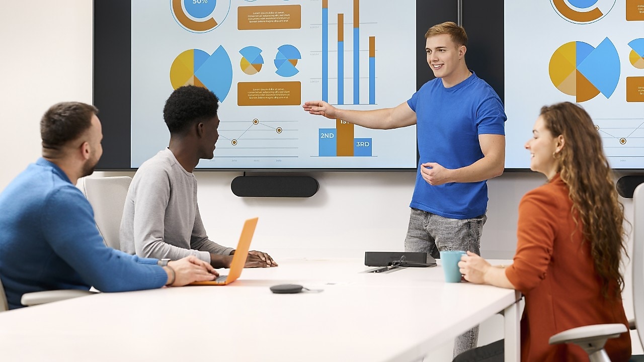 A group of employees, around four of them are in a meeting where one employee is standing and presenting some graphs to the others.