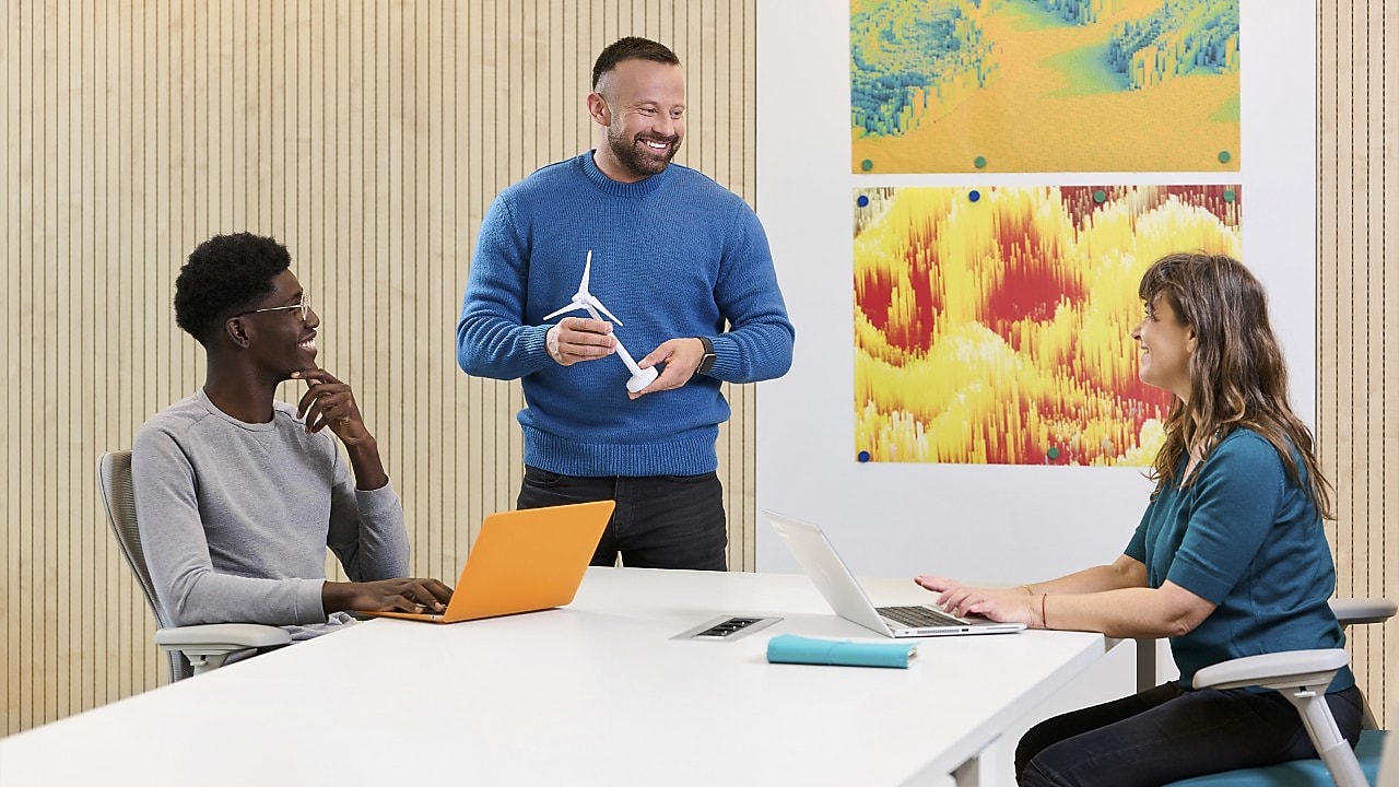 Three employees in a group having a meeting and discussing about work. One employee who is standing has a model/prototype of a windmill in his hand.