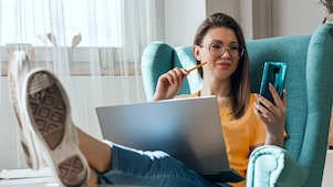 Woman using mobile while looking casually into her mobile phone with a smile; and a laptop is on her lap. She is sitting relaxed on a comfortable chair.