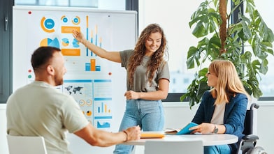 Woman is standing and pointing to a board with colorful charts, while a man and a woman on a wheelchair sit at a round table with notebooks.