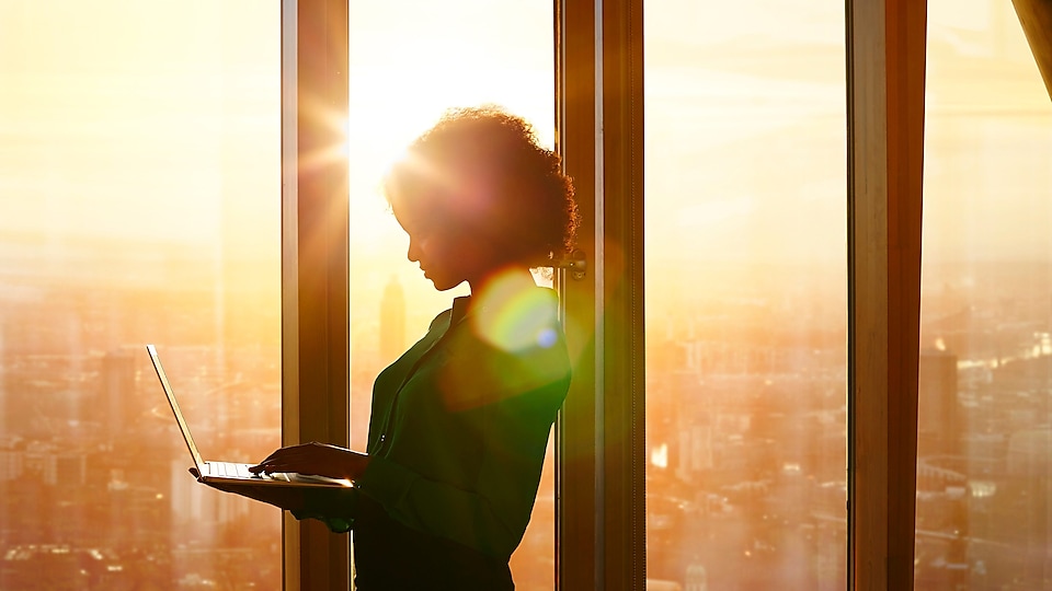 Kobieta stojąca z laptopem na tle zachodzącego słońca za oknami budynku