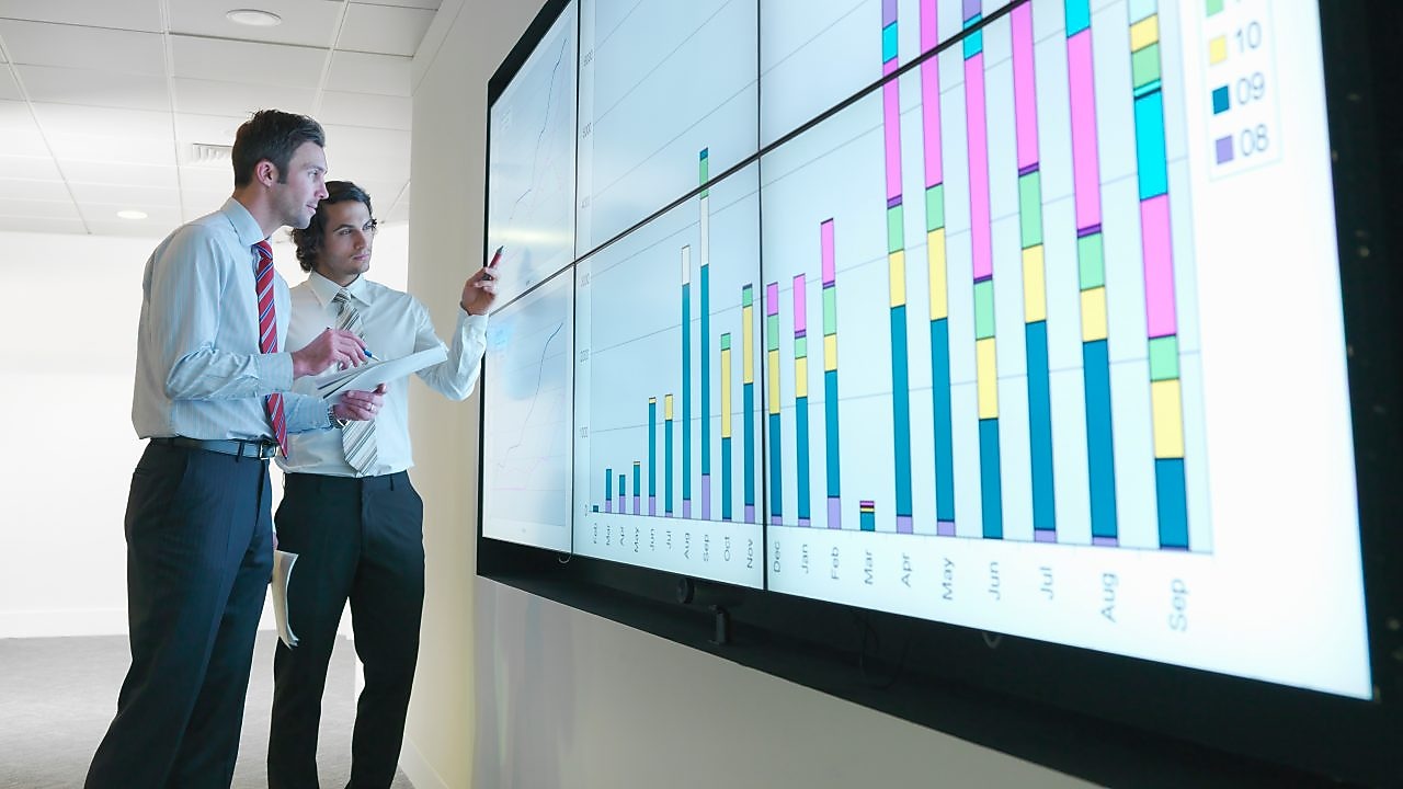 Business men analyse reports displayed on a large digital screen