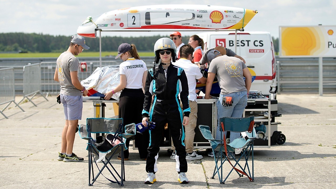 studentka w kombinezonie wyścigowym i kasku stoi na tle naprawianego bolidu 