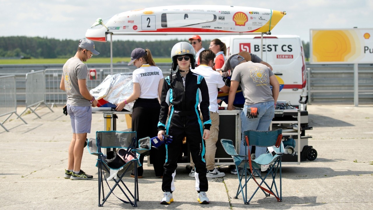 studentka w kombinezonie wyścigowym i kasku stoi na tle naprawianego bolidu 