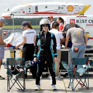 studentka w kombinezonie wyścigowym i kasku stoi na tle naprawianego bolidu 