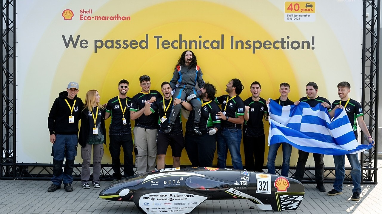 uśmiechnięta grupa studentów z grecką flagą  stroją za czarnym bolidem