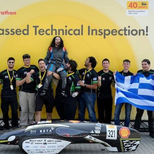 uśmiechnięta grupa studentów z grecką flagą  stroją za czarnym bolidem