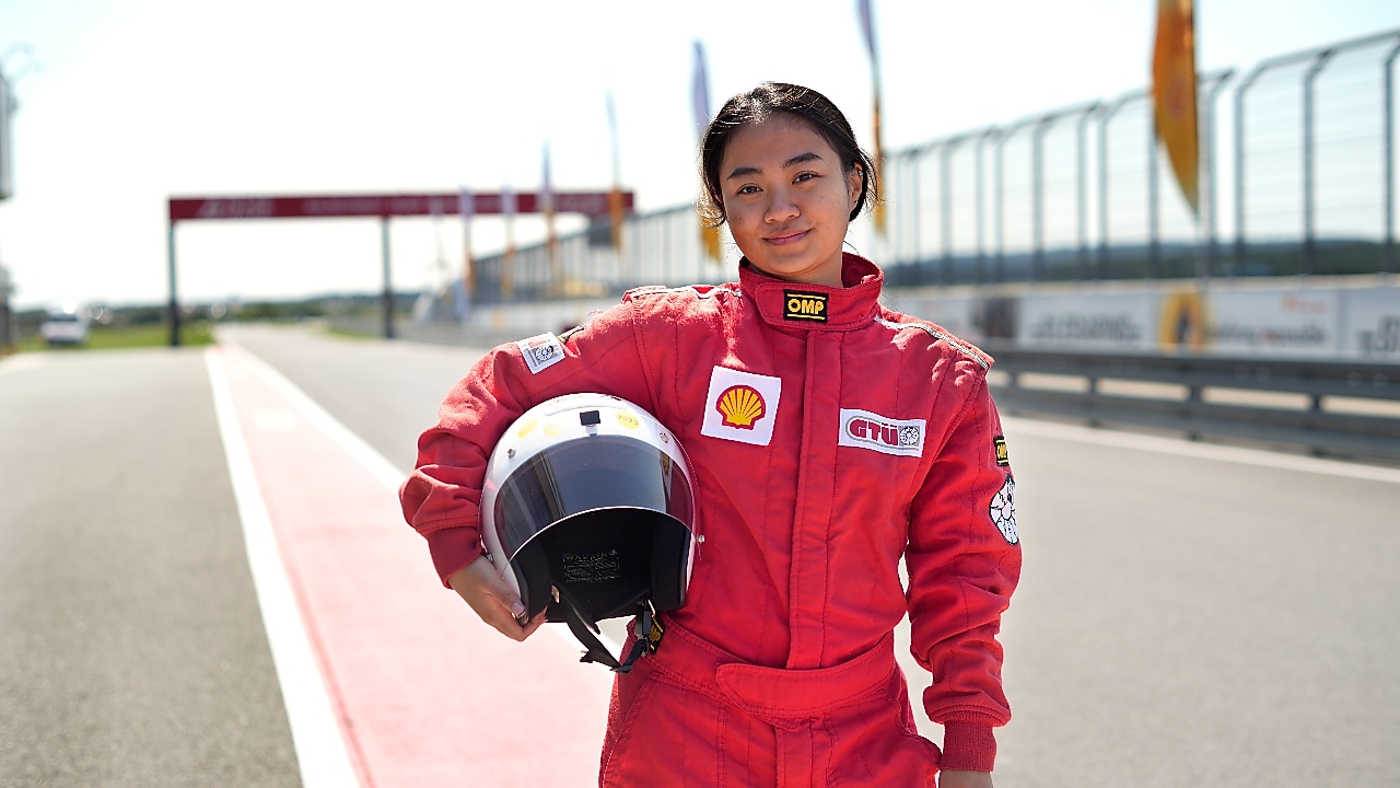 a young person wearing a driving suit and holding a race helmet posing in the pit lane of a race track