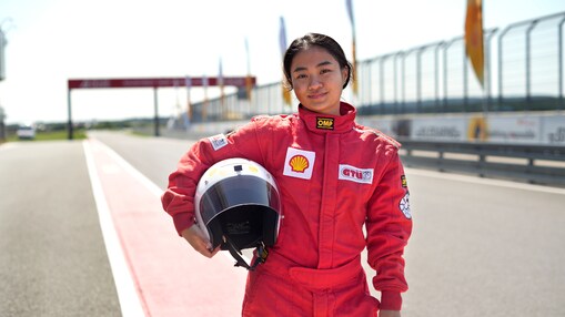 a young person wearing a driving suit and holding a race helmet posing in the pit lane of a race track