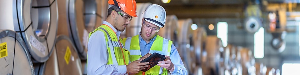 2 mężczyzn w kamizelkach odblaskowych i kaskach ochronnych patrzy na tablet, w tle widać stosy materiałów przemysłowych
