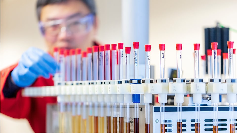 Mężczyzna w czerwonym fartuchu laboratoryjnym i niebieskich rękawiczkach zajmuje się probówkami zawierającymi próbki płynów w laboratorium.