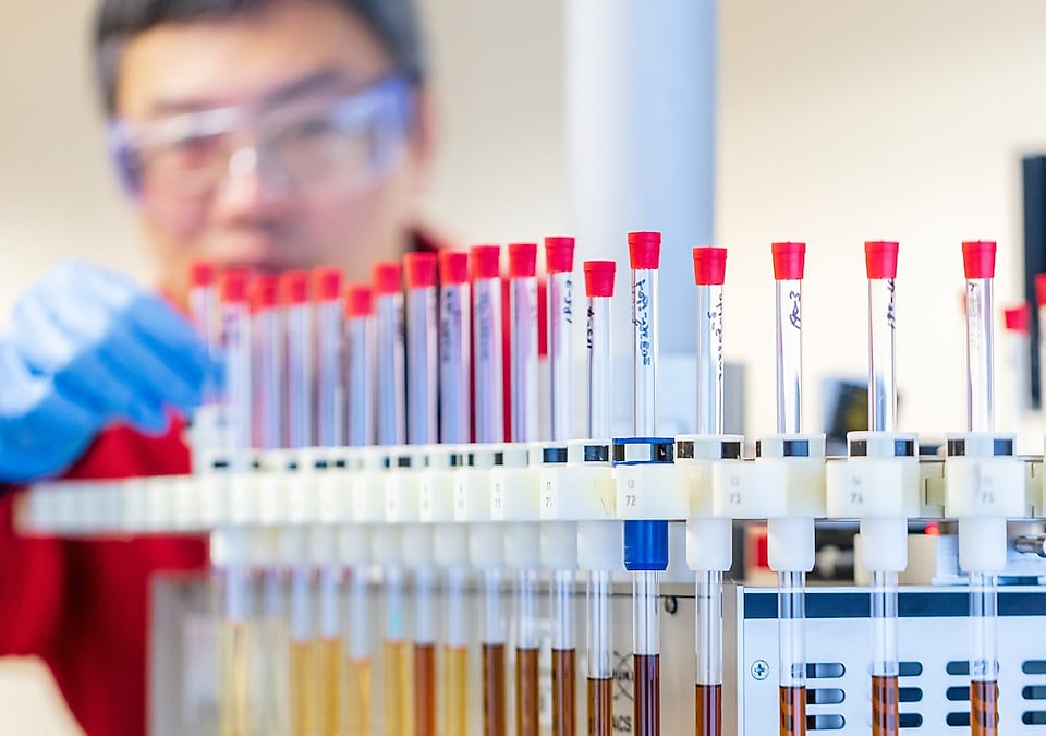 Mężczyzna w czerwonym fartuchu laboratoryjnym i niebieskich rękawiczkach zajmuje się probówkami zawierającymi próbki płynów w laboratorium.