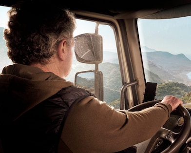 A truck driver steering along a coastal road with a scenic view of mountains and the sea.