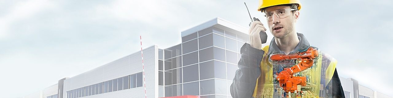 mężczyzna w żółtym kasku rozmawiający przez krótkofalówkę na tle dużego budynku