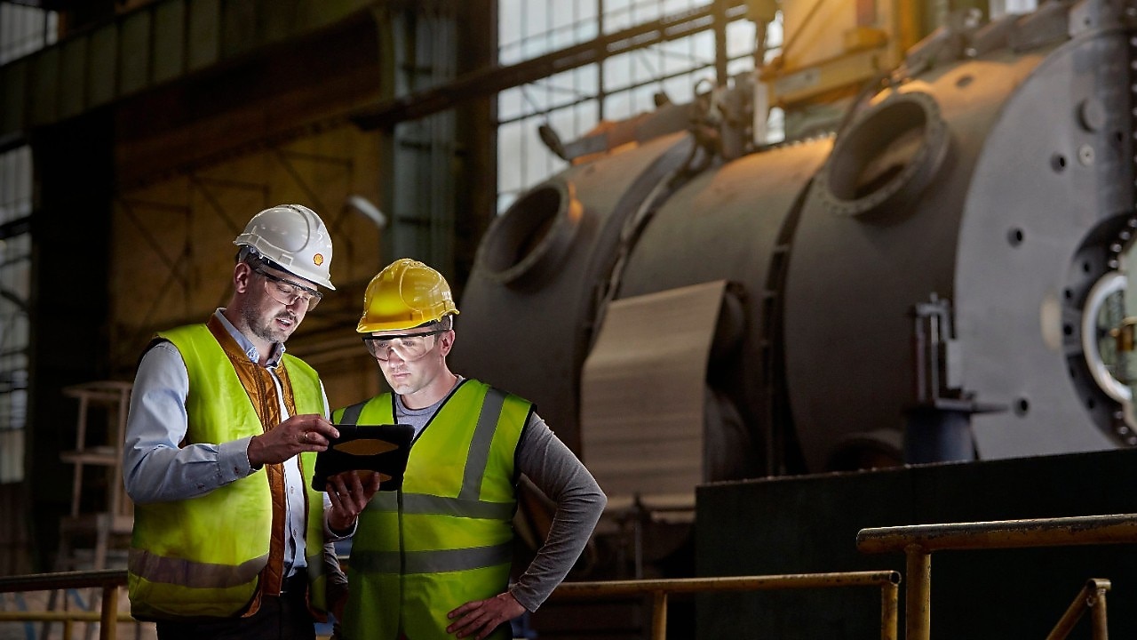 Dwóch mężczyzn w kamizelkach odblaskowych, kaskach i okularach ochronnych na pierwszym planie, w tle duże maszyny przemysłowe