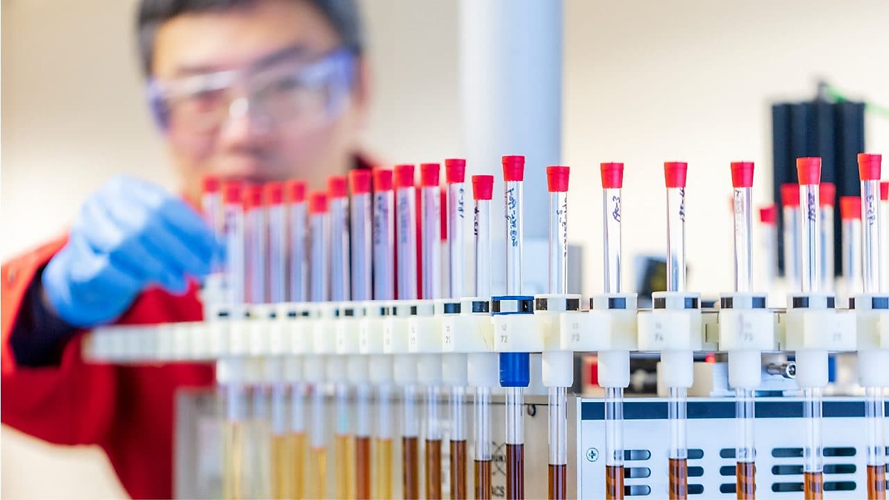 Mężczyzna w czerwonym fartuchu laboratoryjnym i niebieskich rękawiczkach zajmuje się probówkami zawierającymi próbki płynów w laboratorium.