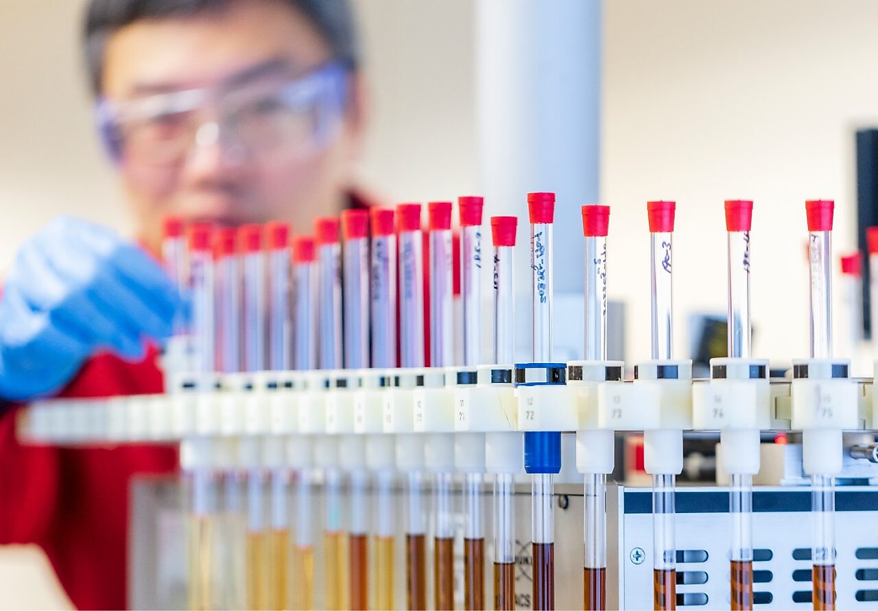 Mężczyzna w czerwonym fartuchu laboratoryjnym i niebieskich rękawiczkach zajmuje się probówkami zawierającymi próbki płynów w laboratorium.