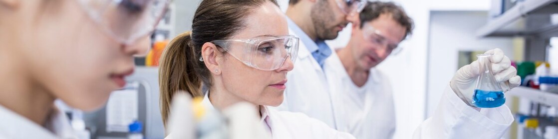 Scientists in a lab with white lab coats and safety glasses handle samples