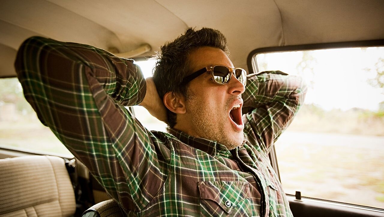 a man yawns in a car, indicating that he needs sleep