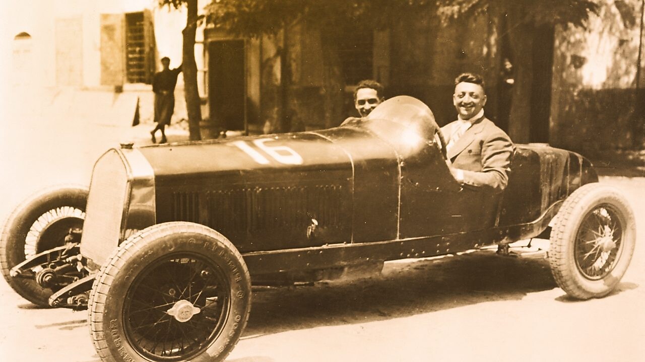 Czarno-białe zdjęcie Enzo Ferrari siedzącego w jednym z pierwszych samochodów Ferrari