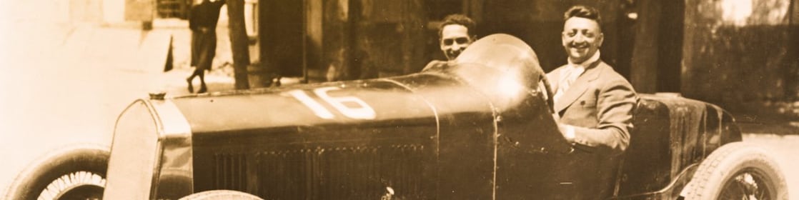 Czarno-białe zdjęcie Enzo Ferrari siedzącego w jednym z pierwszych samochodów Ferrari