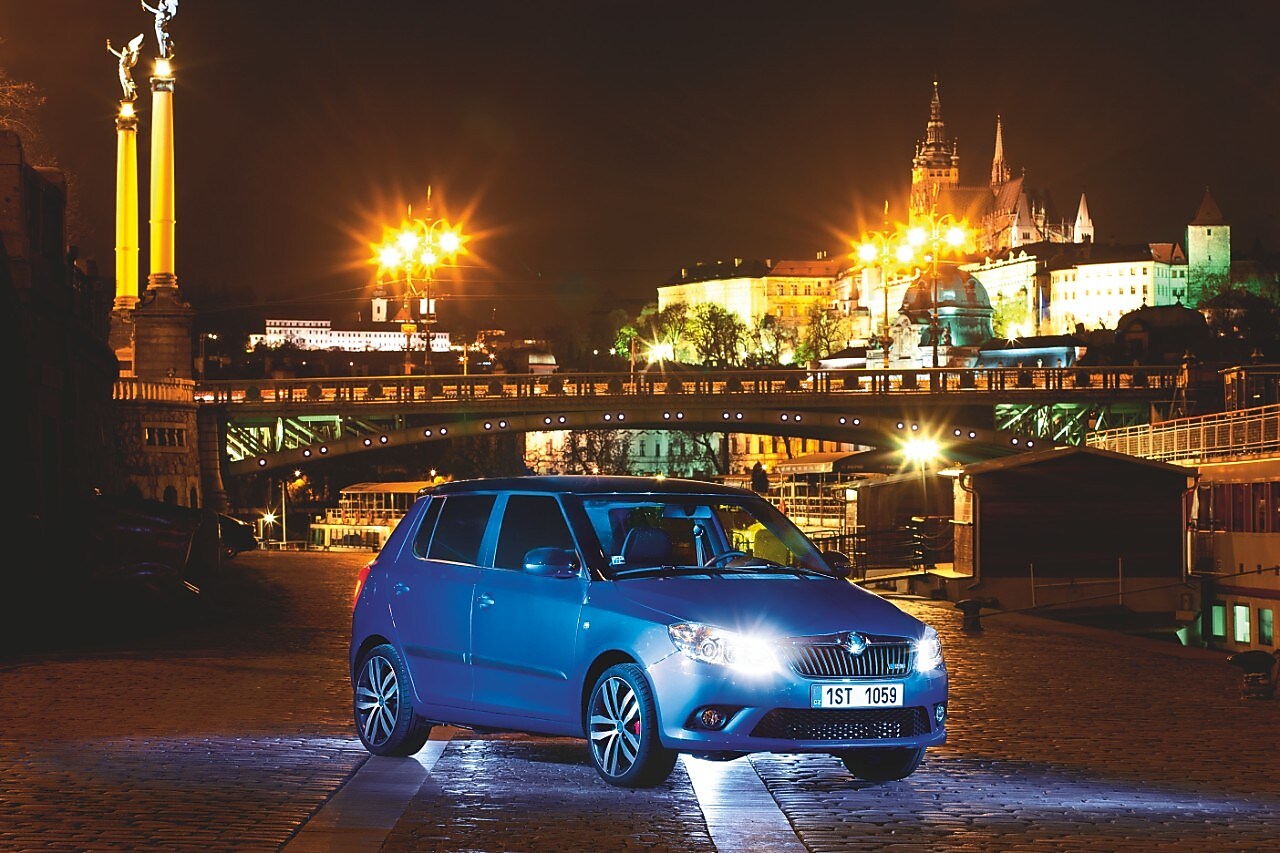 Niebieski samochód zaparkowany nocą na praskiej uliczce, z mostem i zamkiem w tle