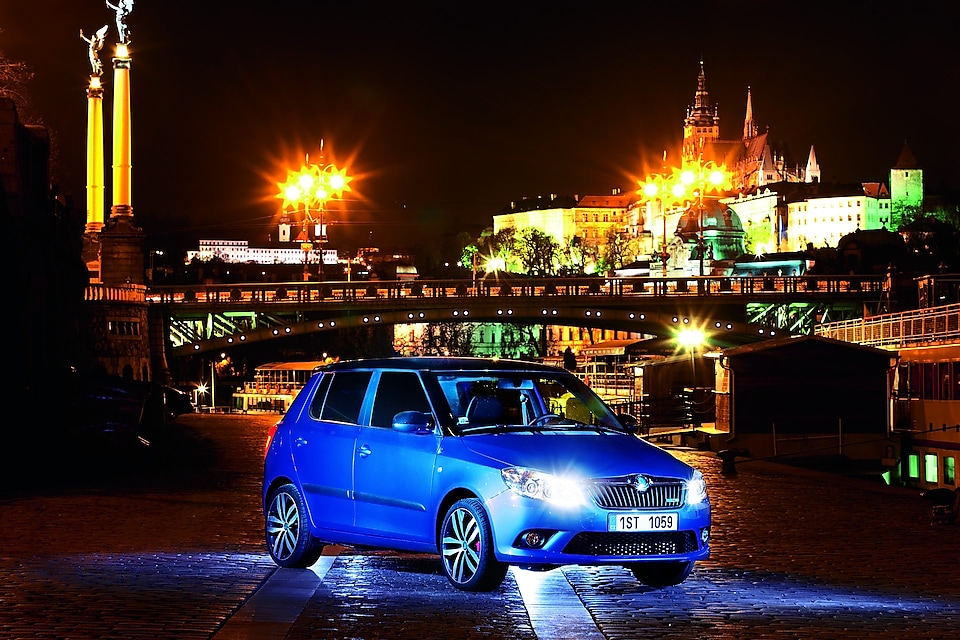 Niebieski samochód zaparkowany nocą na praskiej uliczce, z mostem i zamkiem w tle