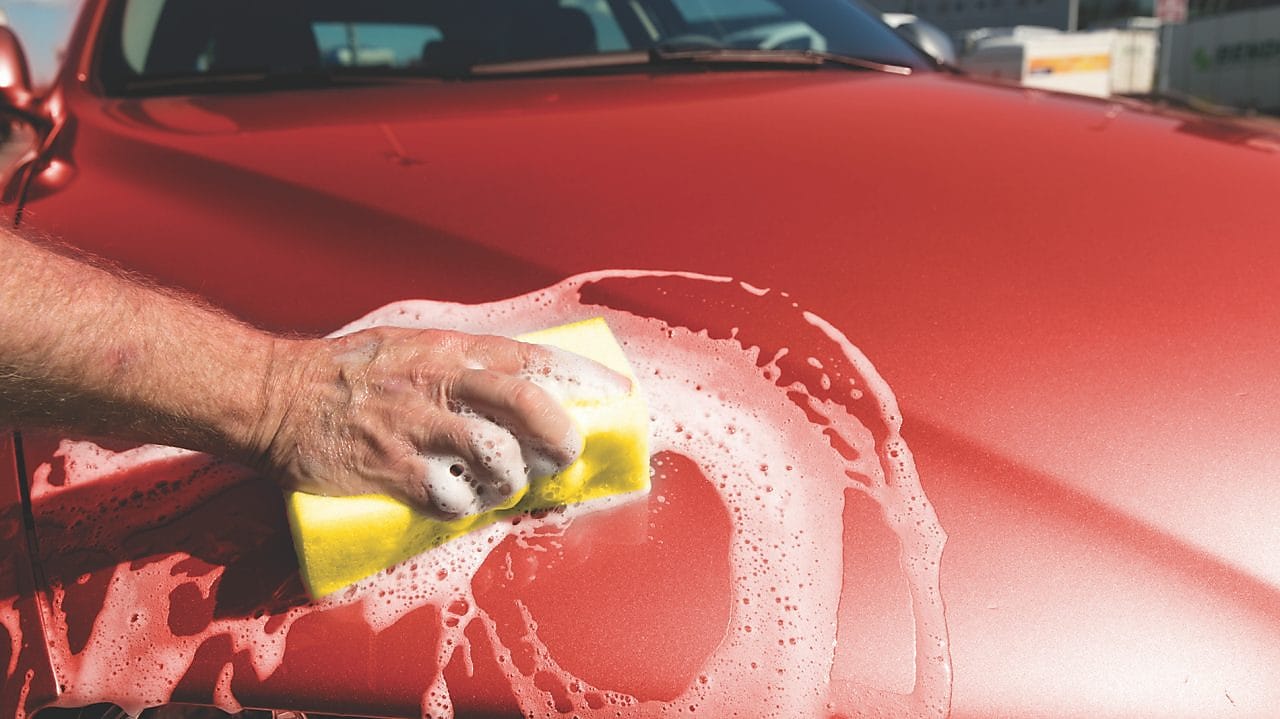 Ręka mężczyczny myjąca maskę czerwonego samochodu za pomocą gąbki z pianą na karoserii.