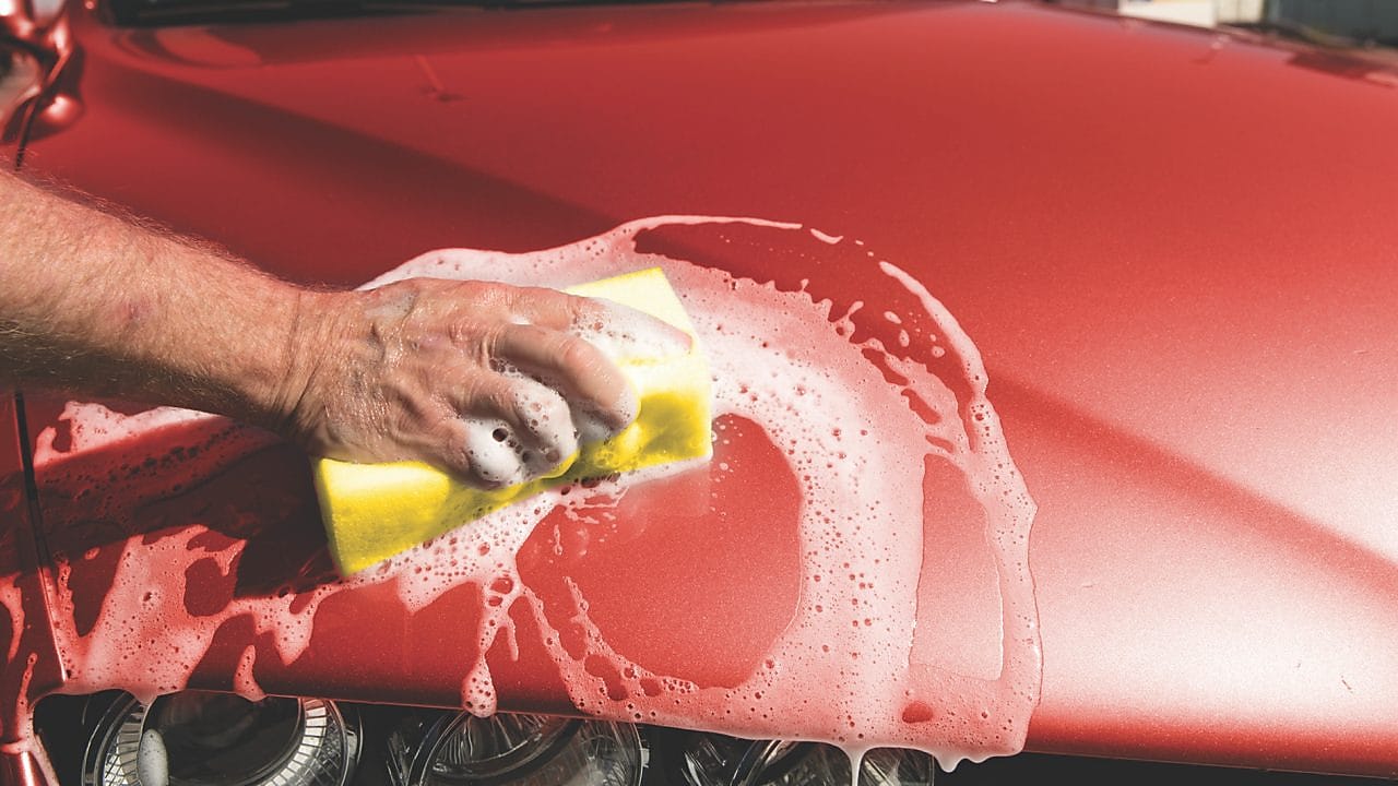 Ręka mężczyczny myjąca maskę czerwonego samochodu za pomocą gąbki z pianą na karoserii.