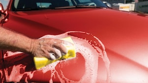 Ręka mężczyczny myjąca maskę czerwonego samochodu za pomocą gąbki z pianą na karoserii.