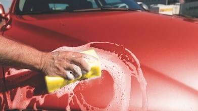 Ręka mężczyczny myjąca maskę czerwonego samochodu za pomocą gąbki z pianą na karoserii.