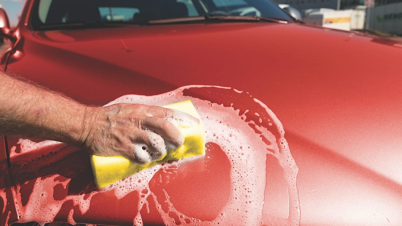 Ręka mężczyczny myjąca maskę czerwonego samochodu za pomocą gąbki z pianą na karoserii.